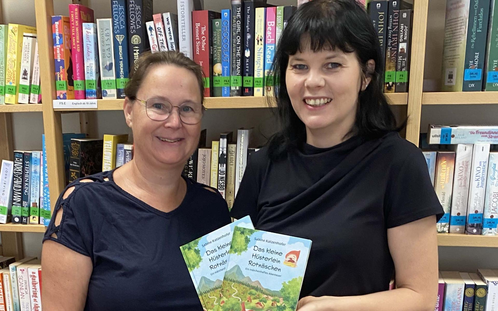 Zwei Frauen vor einer Bücherwand mit einem Kinderbuch in der Hand