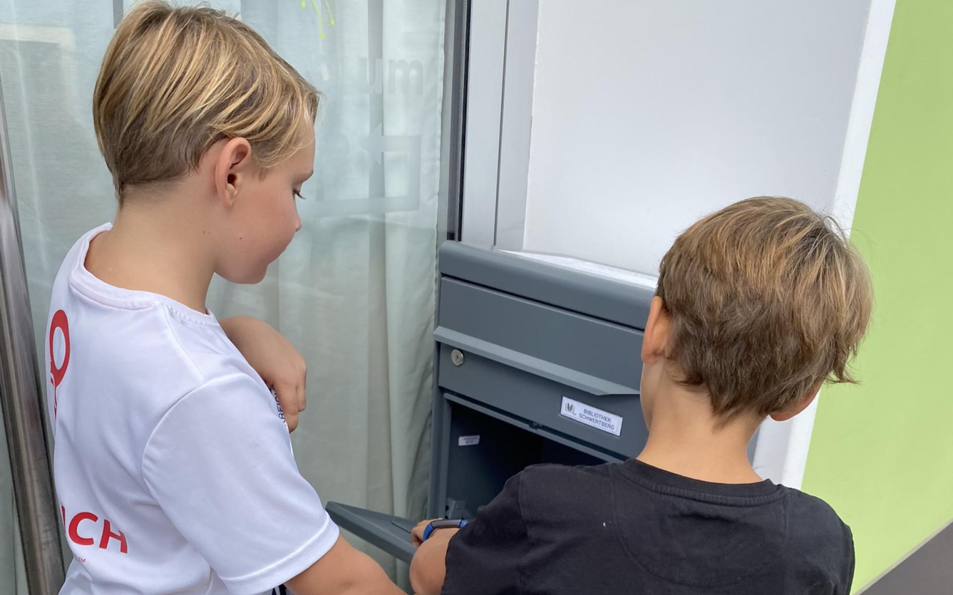 Zwei Kinder geben ihre Bücher in einem Postkasten zurück